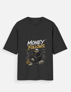A black short-sleeve t-shirt with a graphic print on the front that reads 'MONEY FOLLOWS.' in white and yellow, featuring an illustration of a person in a black hoodie and cap sitting in a chair with stacks of cash and a gold coin.