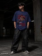 A man wearing a navy blue graphic t-shirt with a red and white anime-style print, paired with black cargo pants and sneakers, standing in a dimly lit concrete parking garage. The brand name KUOWEAR is visible in the bottom-left corner.