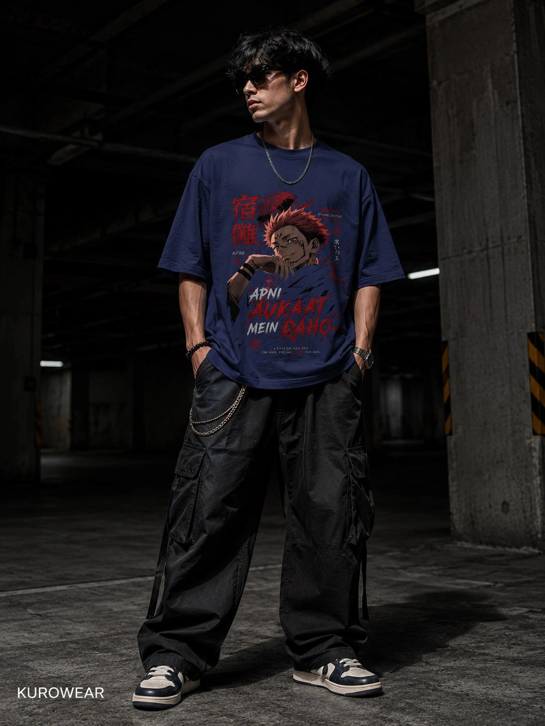 A man wearing a navy blue graphic t-shirt with a red and white anime-style print, paired with black cargo pants and sneakers, standing in a dimly lit concrete parking garage. The brand name KUOWEAR is visible in the bottom-left corner.